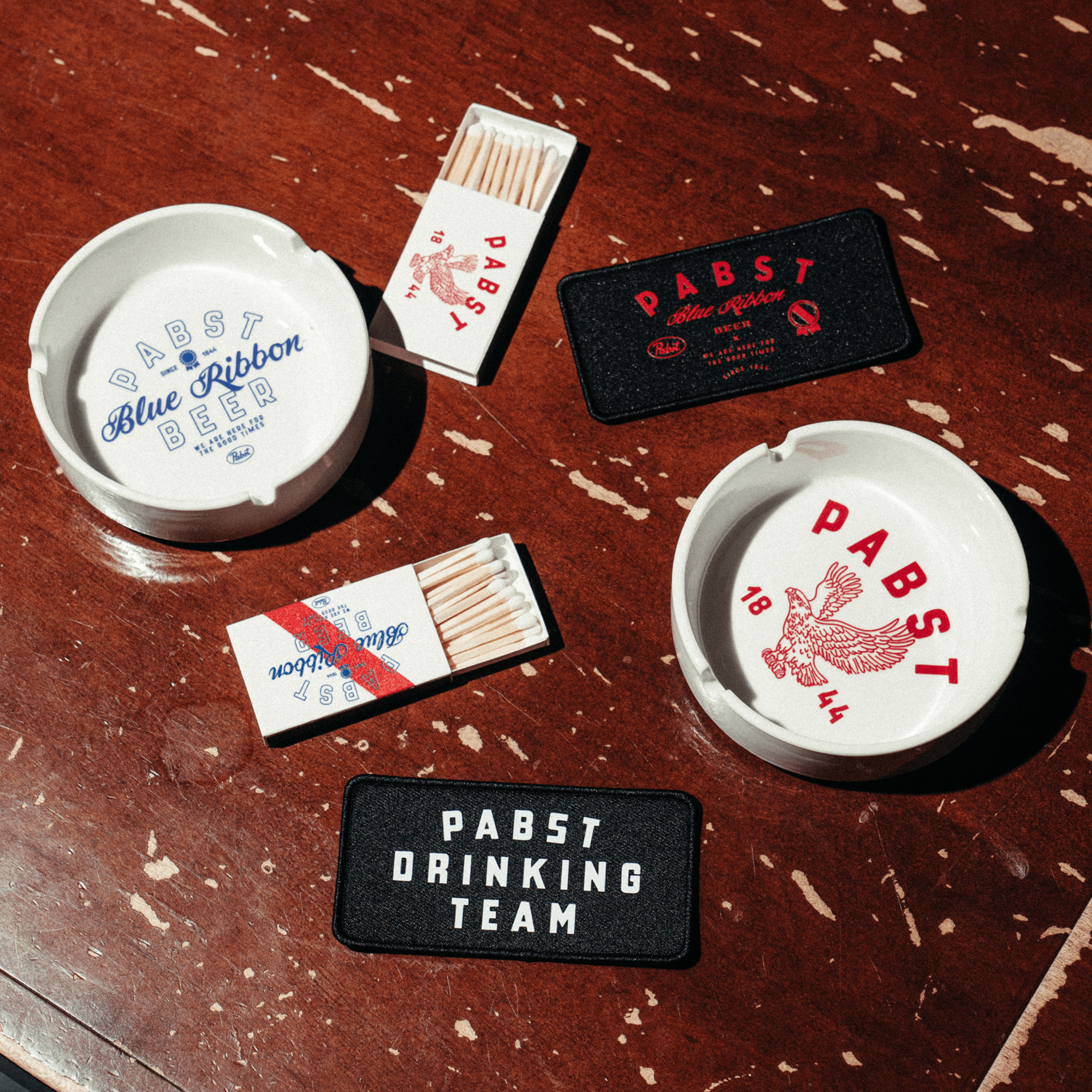 Pabst Blue Ribbon beer-themed items including ashtrays, matches, and patches on a wooden surface.